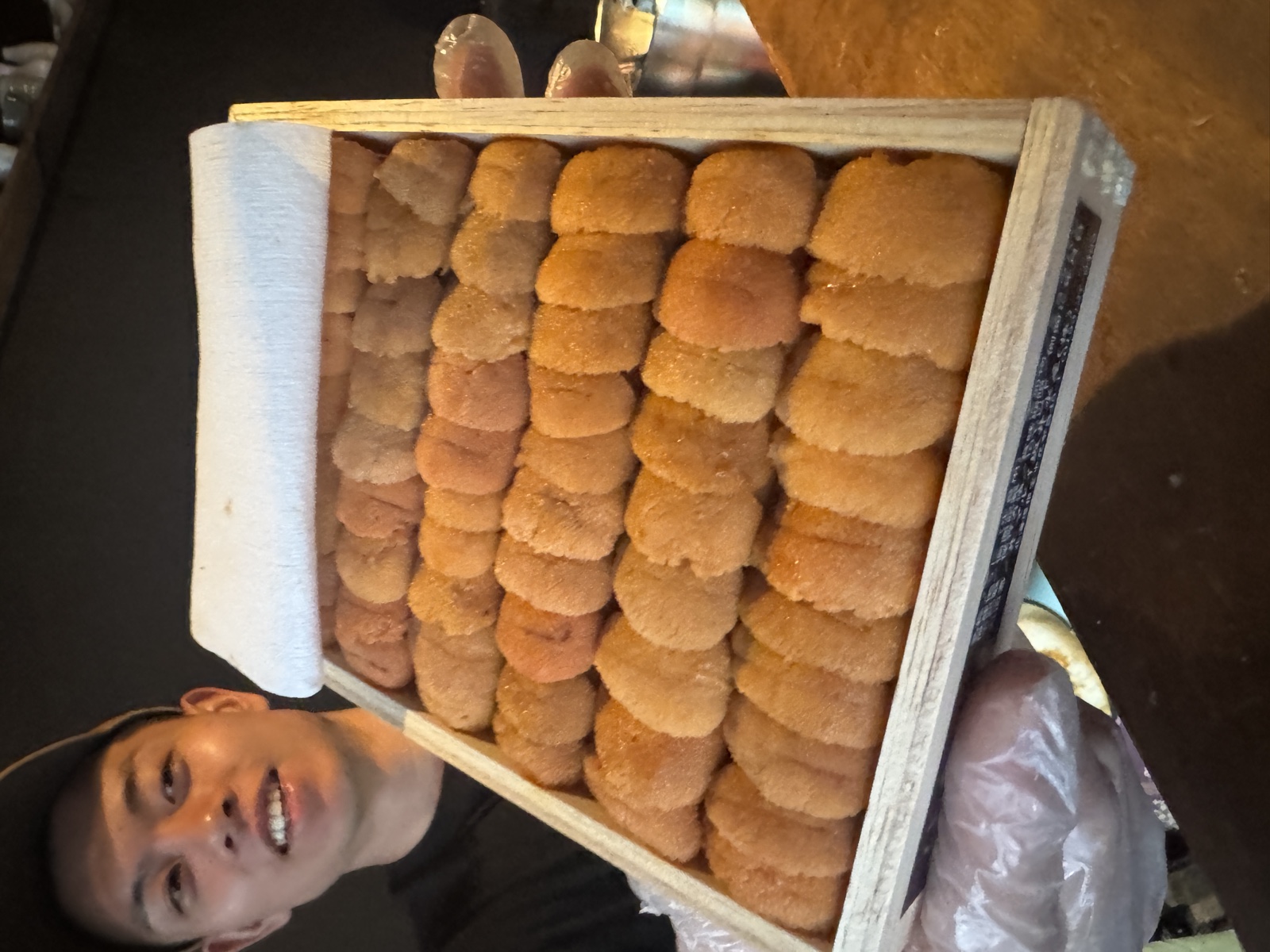 A grinning chef holds a full wooden box of uni lobes toward the camera with gloved hands.