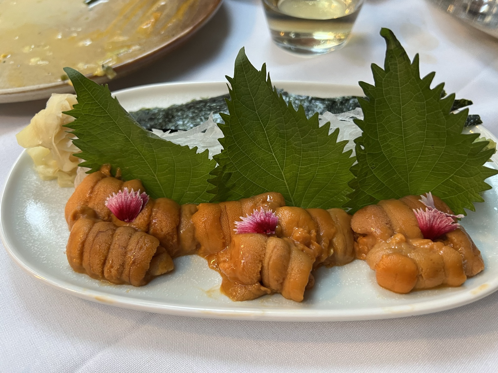 A white plate of pale orange uni lobes laid out with green shiso leaves and pickled flowers, in bright daylight.