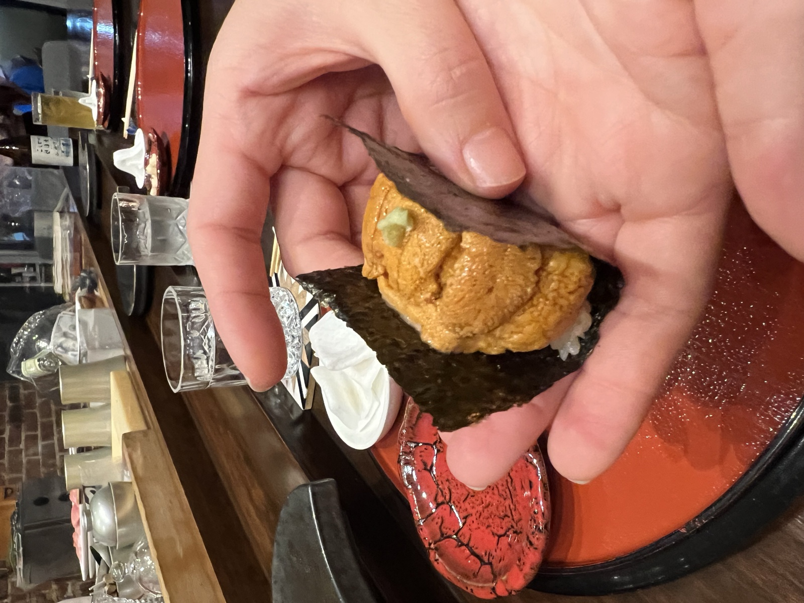 A hand holds a uni temaki, deep orange lobes packed into nori with a purple shiso leaf.
